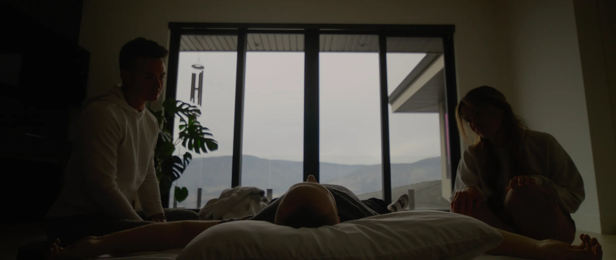 A still from the film. A room with floor-to-ceiling glass over the lake and the mountains behind it, and two facilitators seated on either side of someone lying on a mattress on the floor.