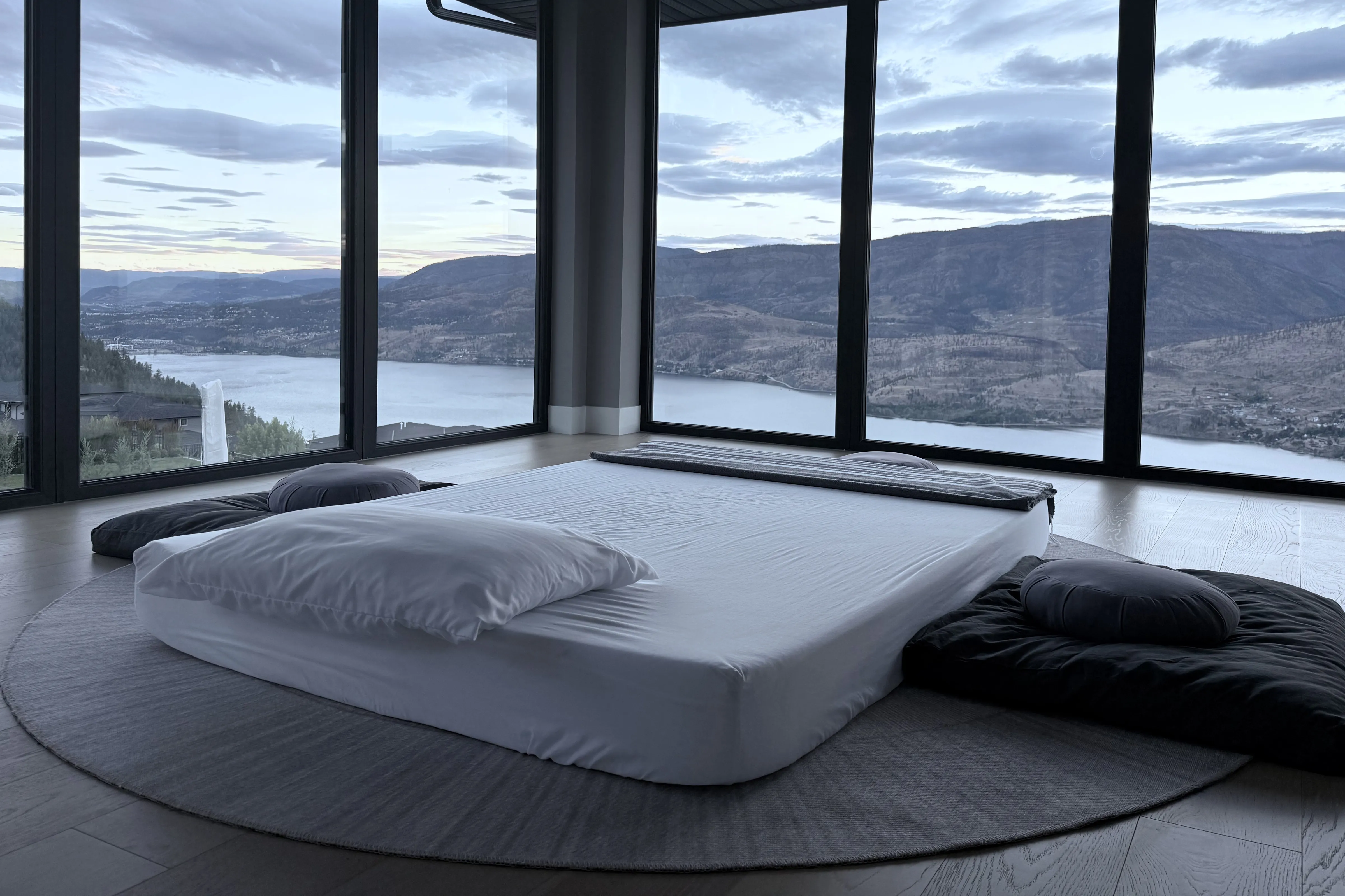 An empty room with corner windows overlooking Okanagan Lake at dusk. A low bed, made, with a floor cushion on either side.
